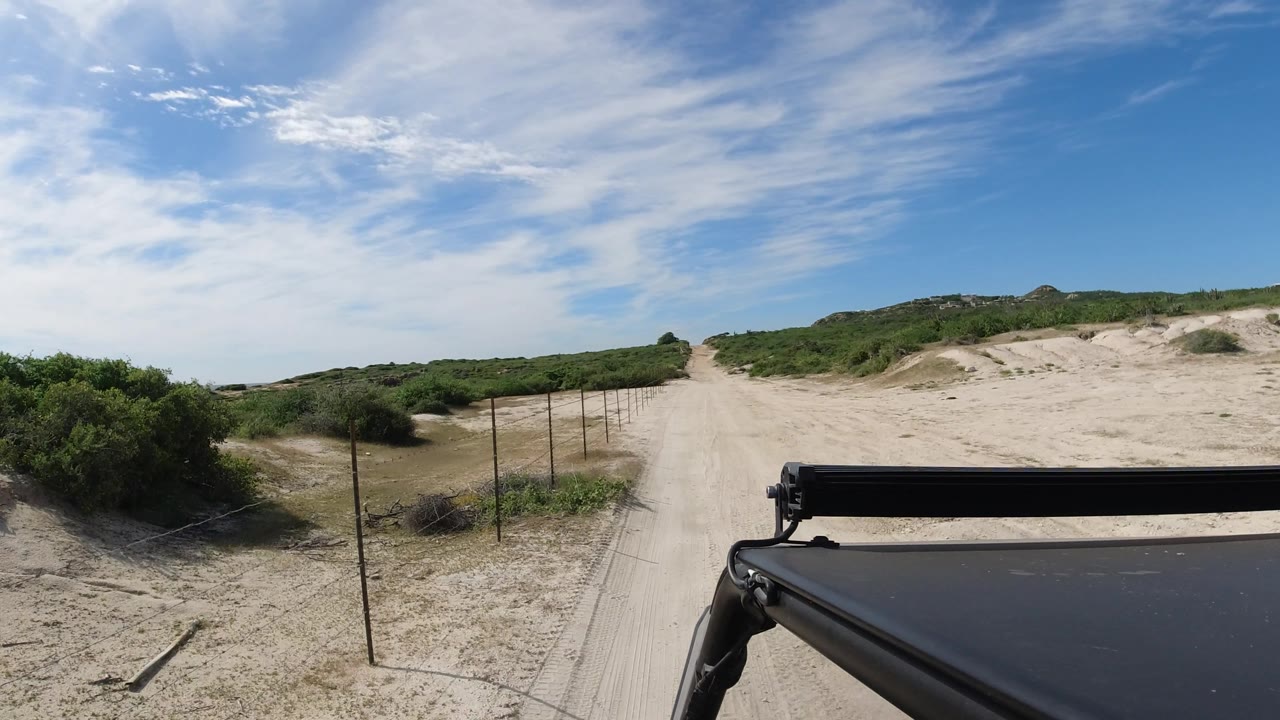 Desert road and dunes with bushes, fence posts, and black vehicle frame.