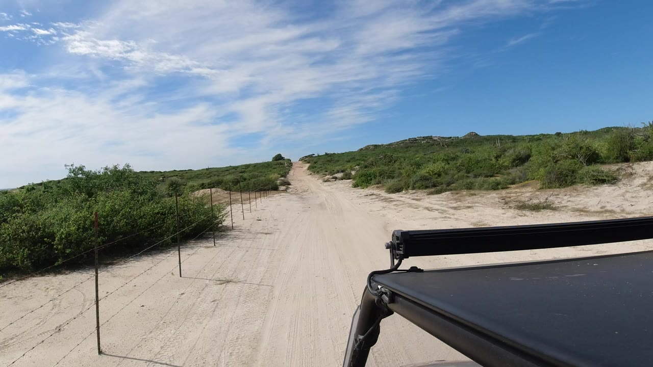 Los Cabos sandy desert road ahead with low green shrubs, blue sky, wispy clouds, a black vehicle roof edge in the foreground, and no visible people.
