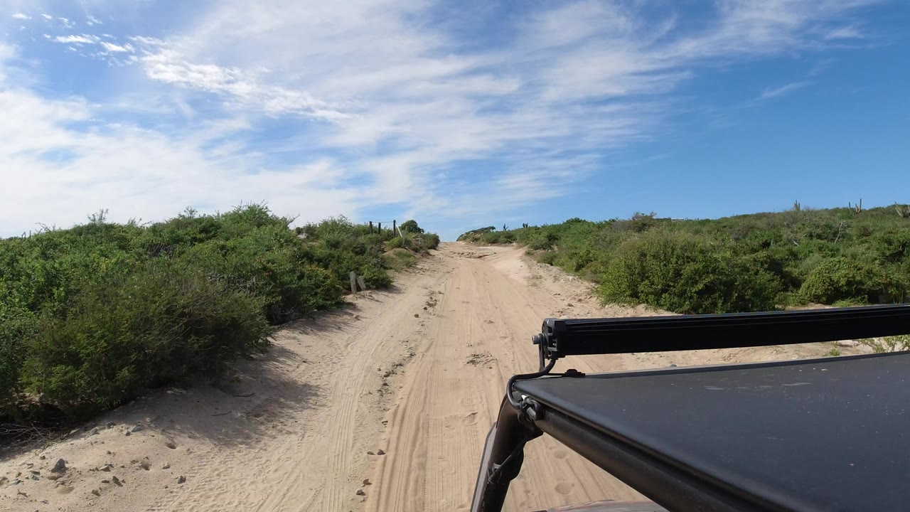 A Los Cabos sandy road continues through dense green scrub, with the vehicle roof edge low in frame and no visible people along the route.