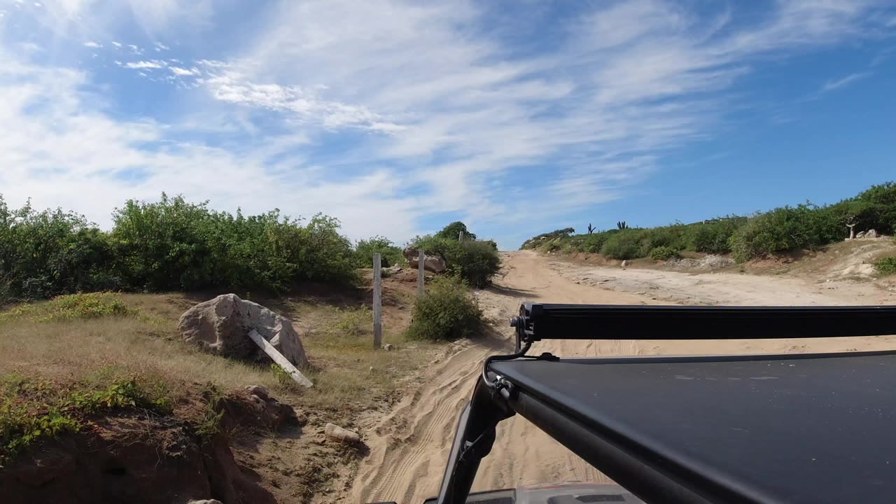 Opening frame of a sandy Los Cabos road bordered by low green scrub, rocks, and wooden posts, seen over a black vehicle roof bar with no visible people.
