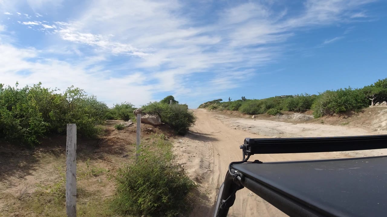 Sandy road curving past posts, bushes, and a vehicle corner.