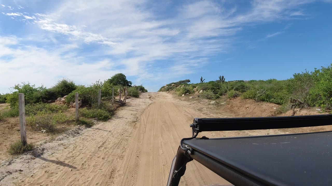 The vehicle viewpoint continues down a sandy Los Cabos road, with low green brush on both sides, fence posts to the left, and no visible people.