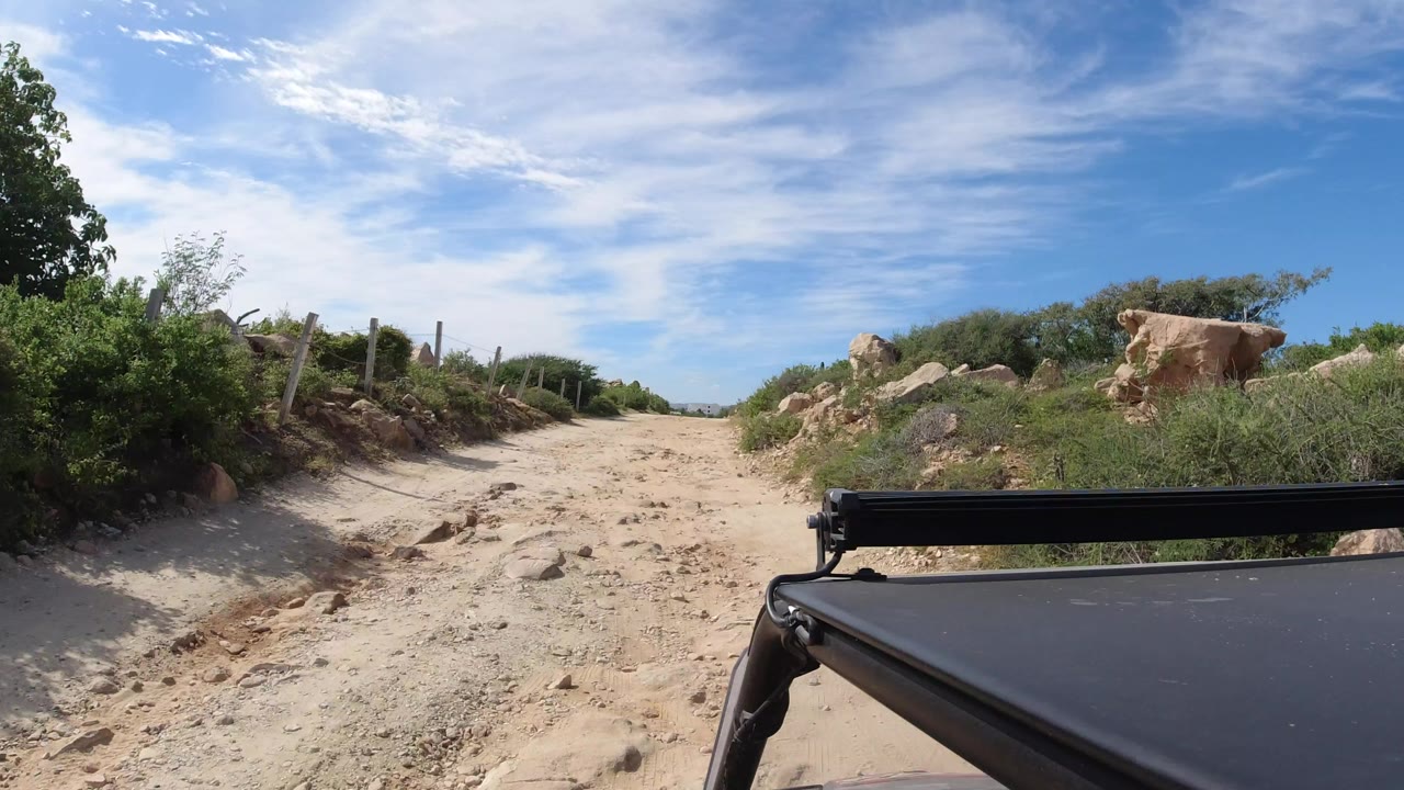 Sandy road bordered by brush and rocks with a black vehicle edge in the foreground.