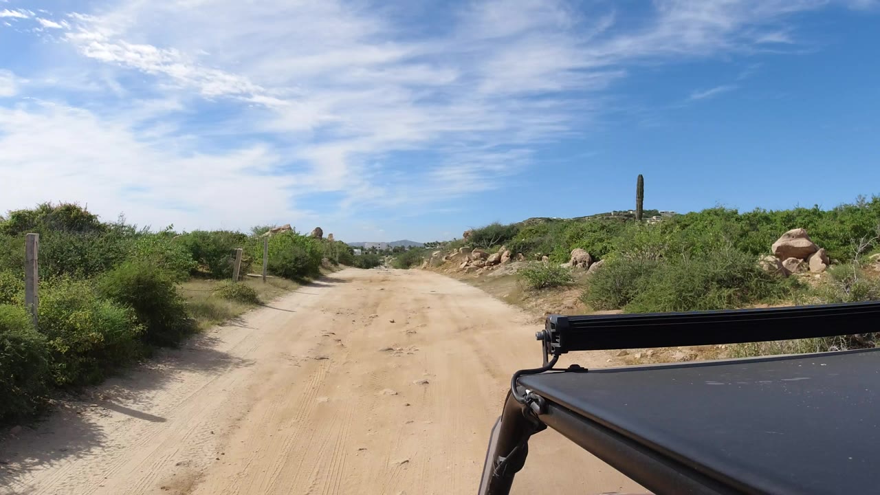 Start of the Los Cabos, Mexico clip looking down a sandy road bordered by low brush and rocks, with part of the Wrangler visible in the foreground and no visible people.