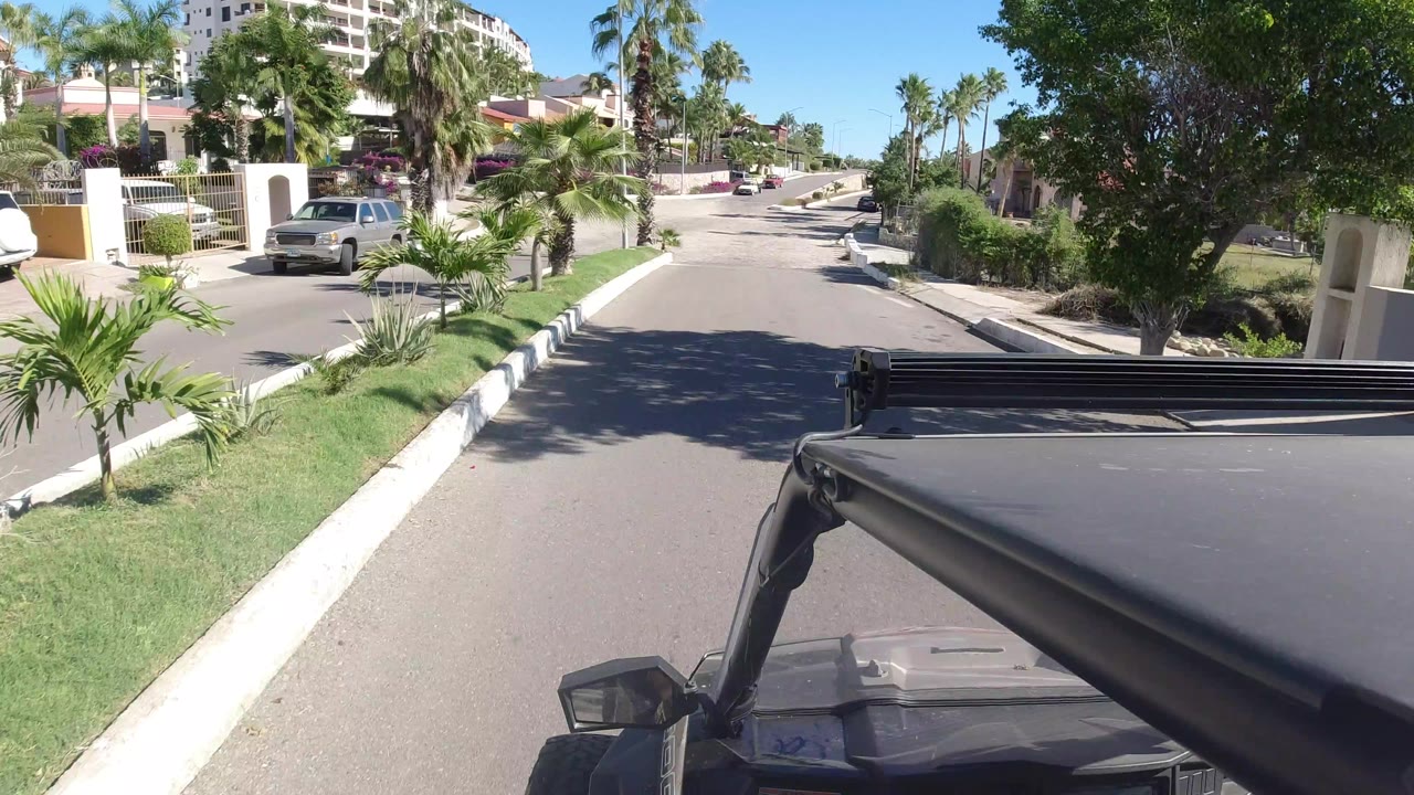 The Wrangler continues along a Los Cabos residential road, passing palms, a grassy verge, white walls, parked cars, and no visible people.