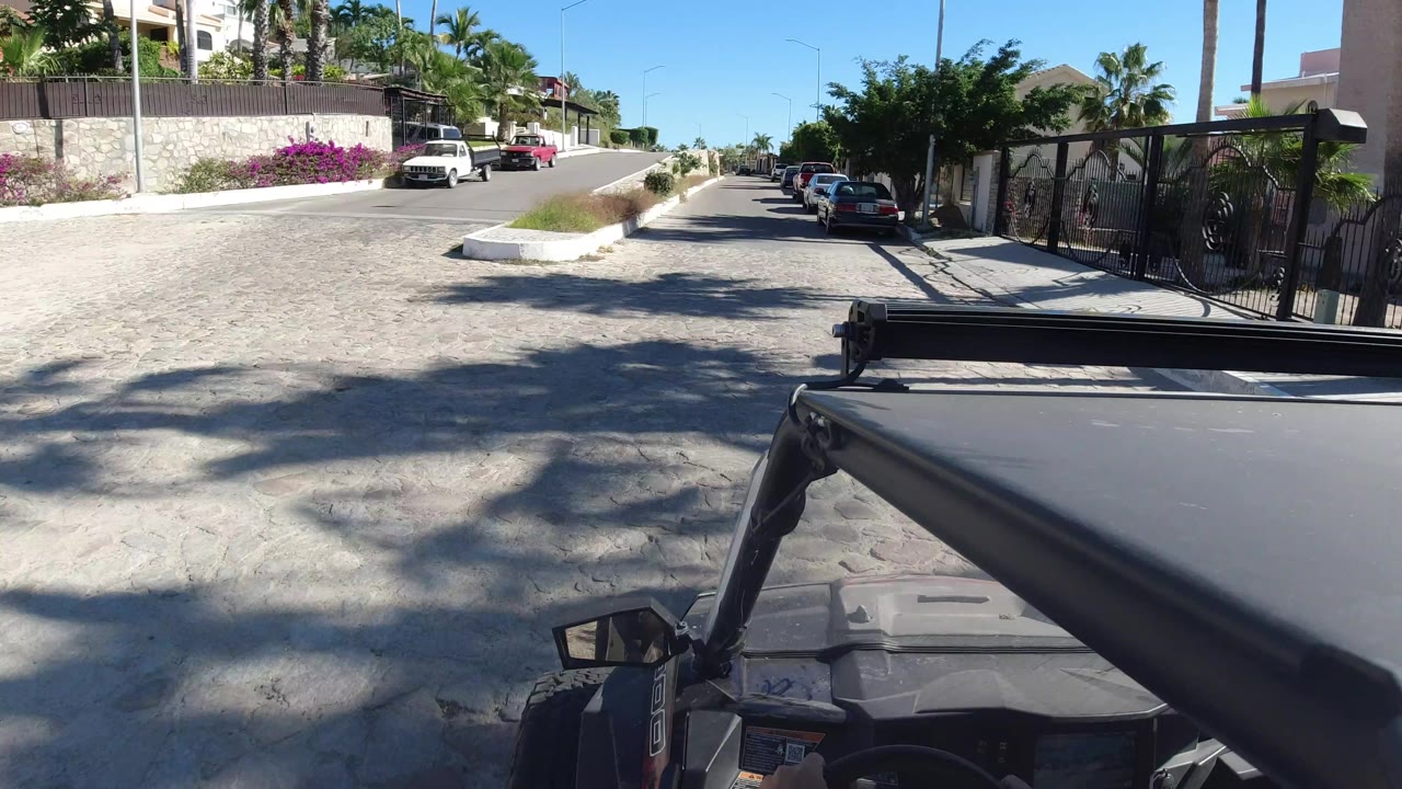 Forward view from a Wrangler in Los Cabos, Mexico, with curbside cars, a white wall, flowering shrubs, gated residences, palm trees, and no visible people.