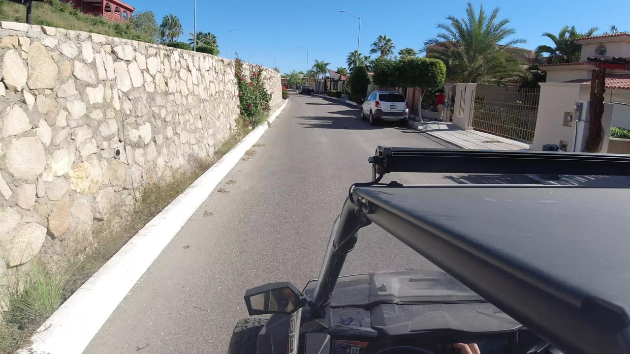 A forward-facing Wrangler view continues down the Los Cabos street, with the stone wall close on the left, a white car ahead, homes to the right, and no visible faces.