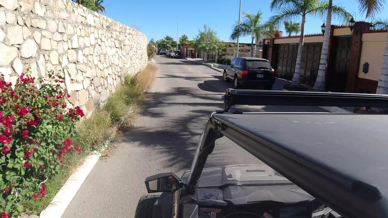 A second view along the same Los Cabos road shows the Jeep foreground, red flowering shrubs against a stone wall, a black parked car, gated homes, palm trees, and bright sun, with no clearly visible people.