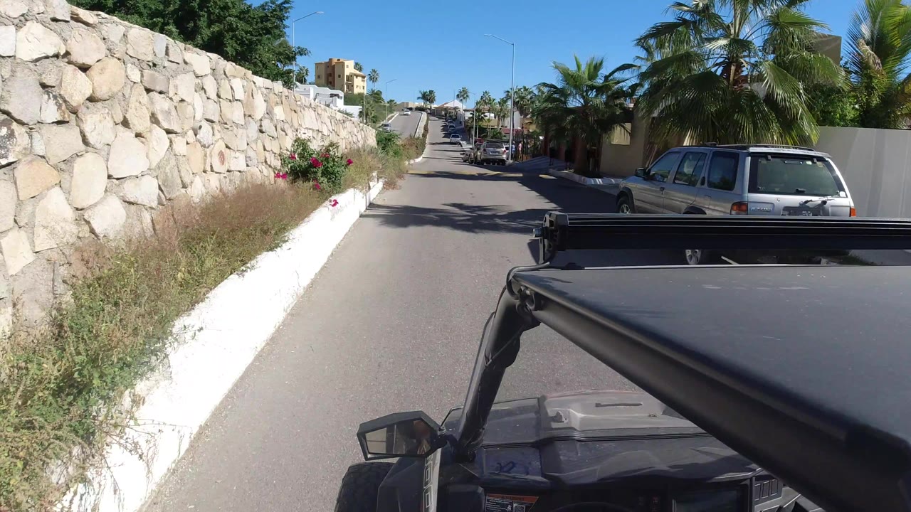 Los Cabos street view from the top of a jeep, with the stone wall closer on the left, a silver parked vehicle on the right, palms, homes, and no visible people.