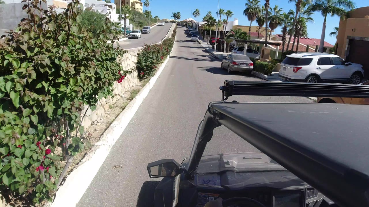 The Los Cabos road remains centered as the Wrangler hood fills the lower foreground, with a stone wall and greenery at left, parked vehicles at right, palms above the houses, and no visible people.