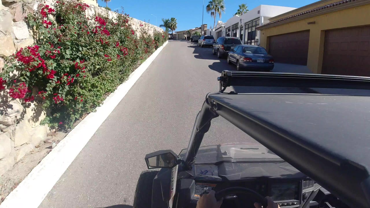 Open vehicle hood on a narrow sunny street beside a stone wall with red flowers.