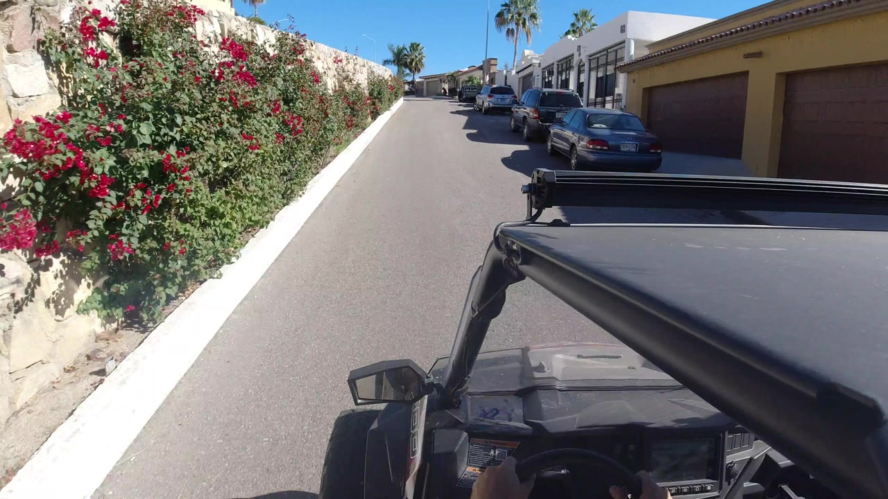 One second into the Los Cabos, Mexico street drive, the vehicle foreground stays low while red flowering shrubs fill the left side and parked cars line the road ahead, with no visible people.