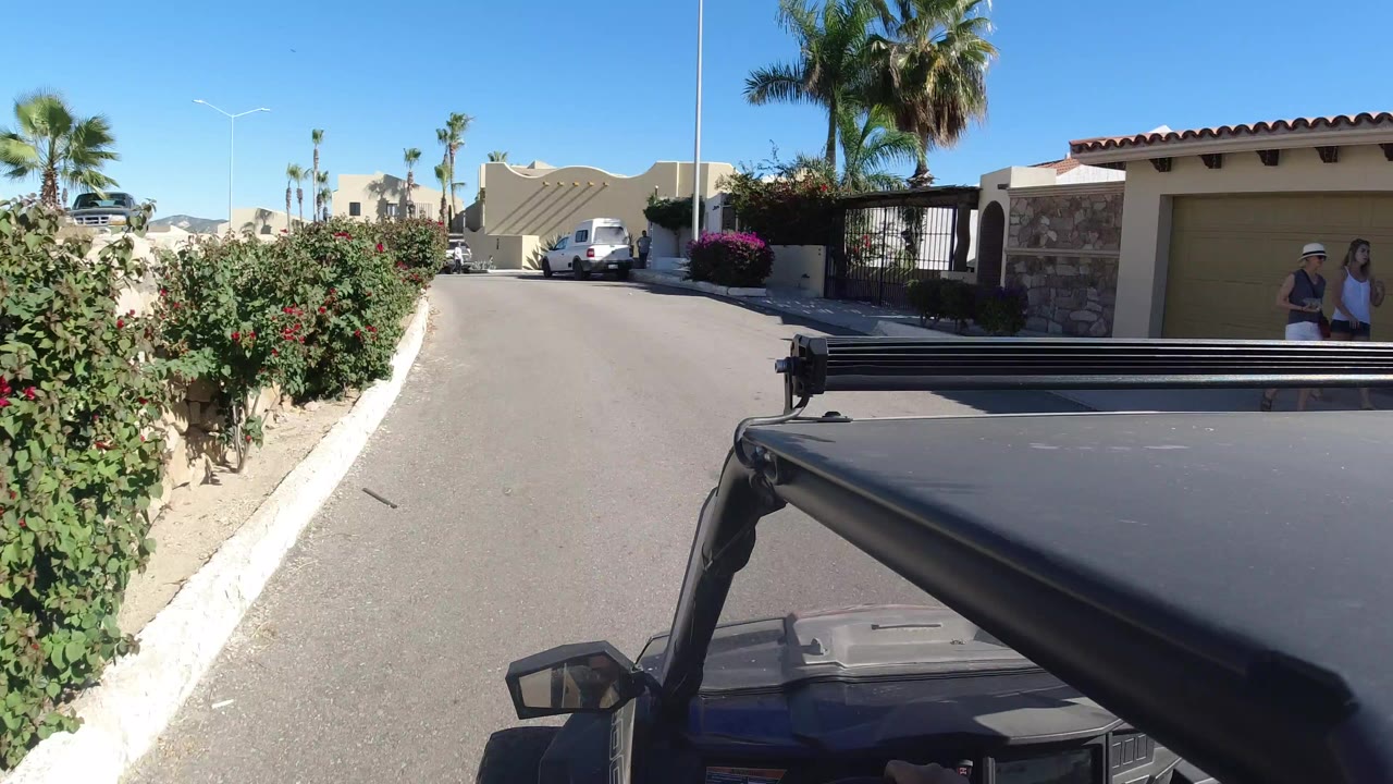 Vehicle view down a sunny residential road with shrubs and beige homes.