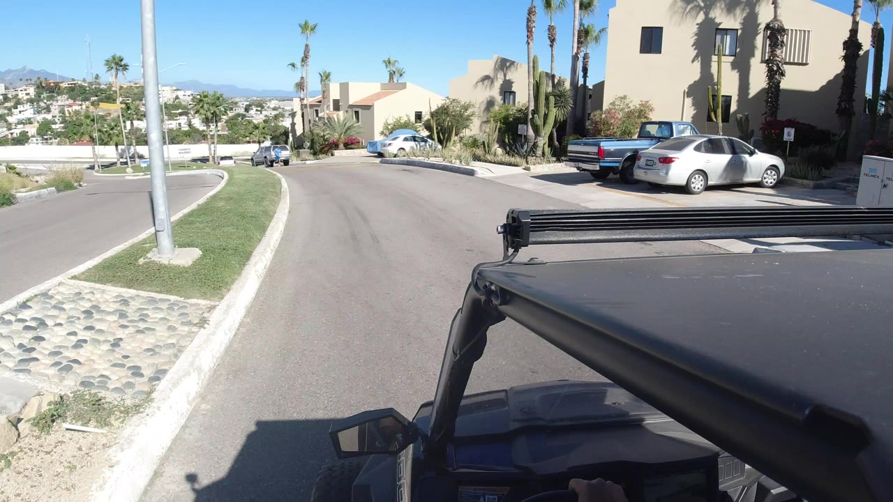 Vehicle roof foreground on a sunny curved street with stucco buildings and palms.