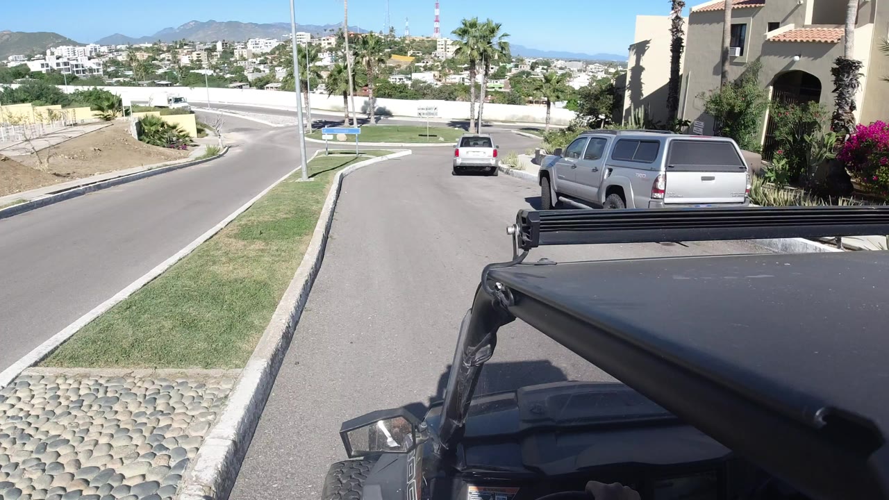 Forward-facing view from a black Wrangler in Los Cabos, Mexico, following a silver SUV past grass strips, palms, homes, and distant hills, with no visible people.