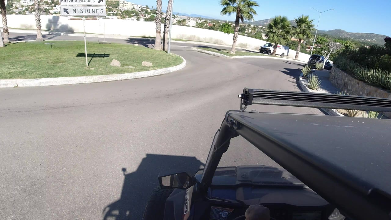 Los Cabos road POV from the Wrangler with the hood and rail in the foreground, a bend ahead, palms along the curb, a white wall, and a white parked van in bright sun, with no visible people.