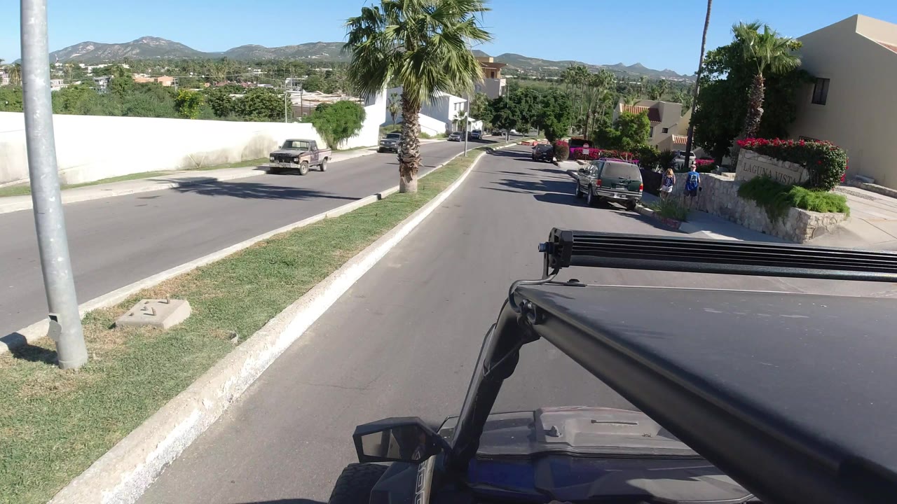 A vehicle-mounted view in Los Cabos looks along the road past a tall palm, parked cars, white stucco buildings, low hills, and no clearly visible people.
