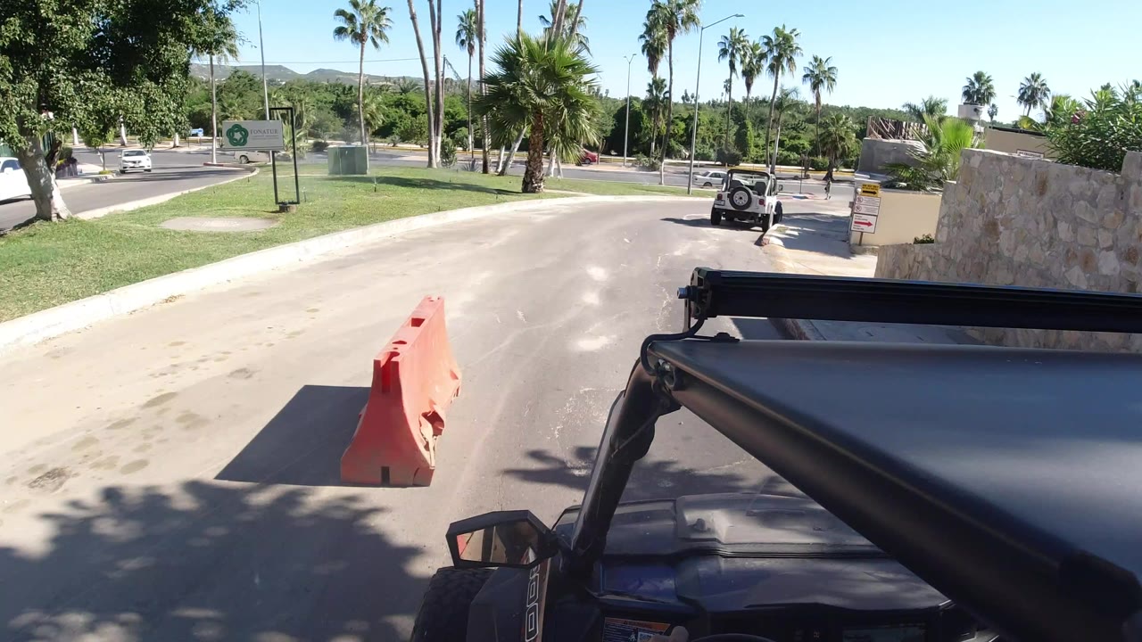 A black Wrangler fills the lower right foreground on a sunny Los Cabos road, with an orange barrier, palm trees, grass, and a distant vehicle ahead, no visible people.