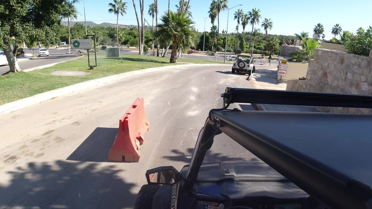 The Los Cabos boulevard remains in view from the Wrangler, with the orange barrier closer to center, palms lining both sides, and a small vehicle ahead, no visible people.
