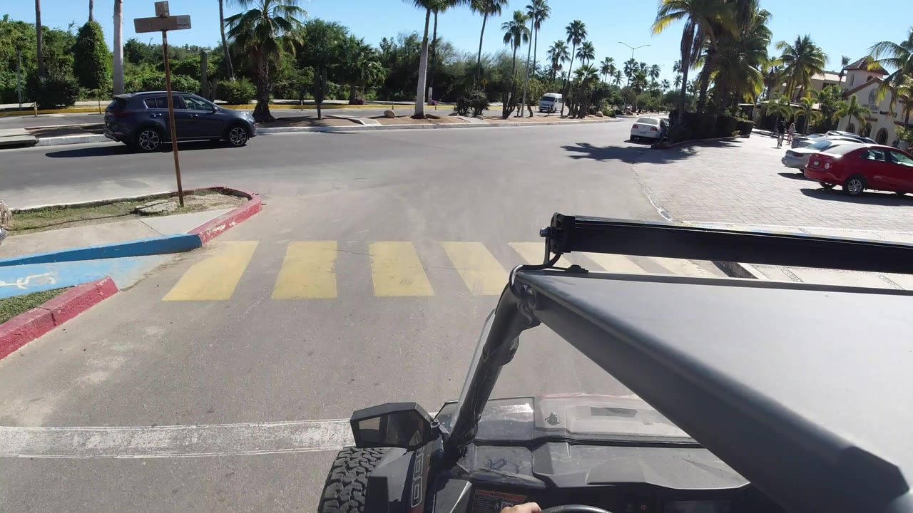 Front passenger view from a Jeep at a Los Cabos road crossing with yellow crosswalk stripes, parked cars, palms, and no visible people.