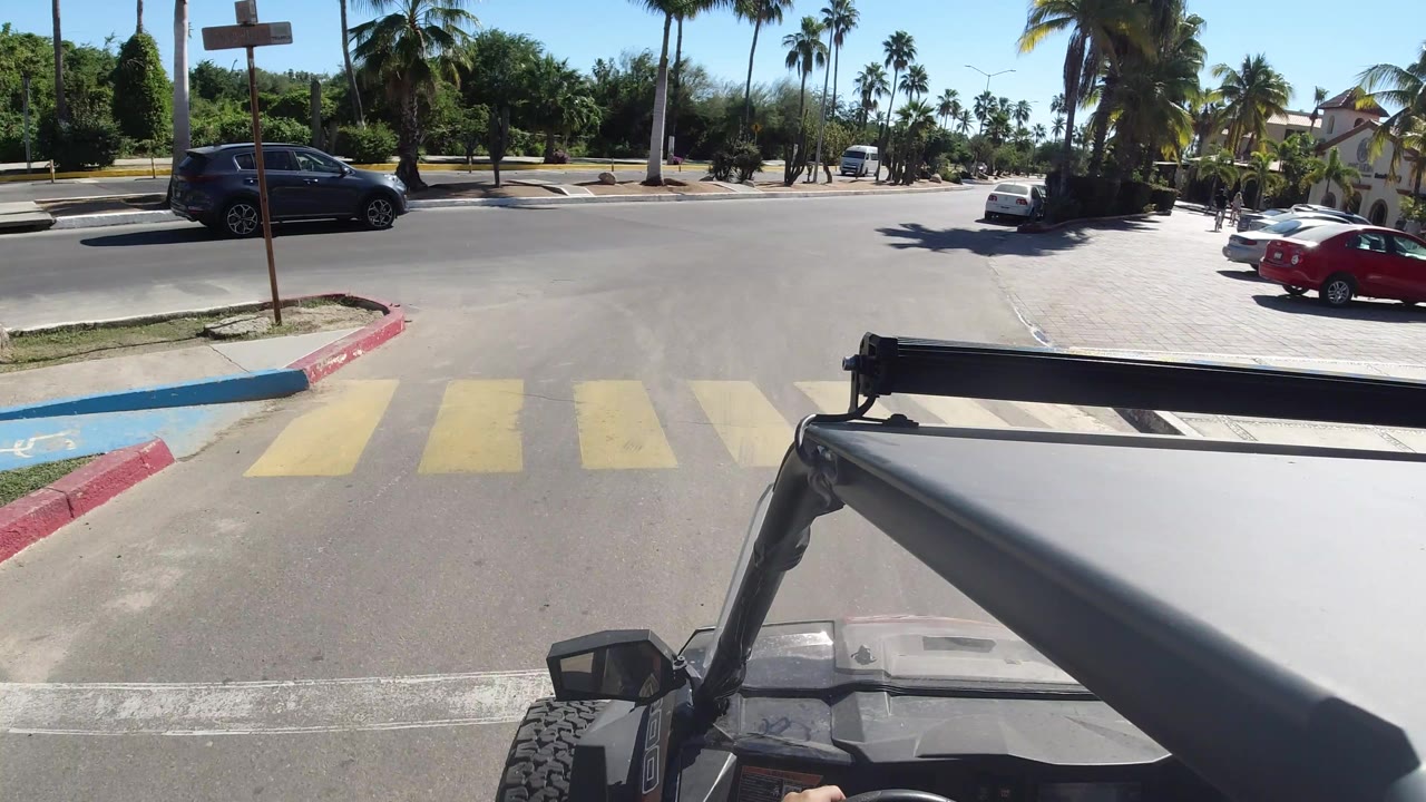 The Jeep view continues along a sunlit Los Cabos street with a red car, dark SUV, curb paint, palm trees, and no visible people.