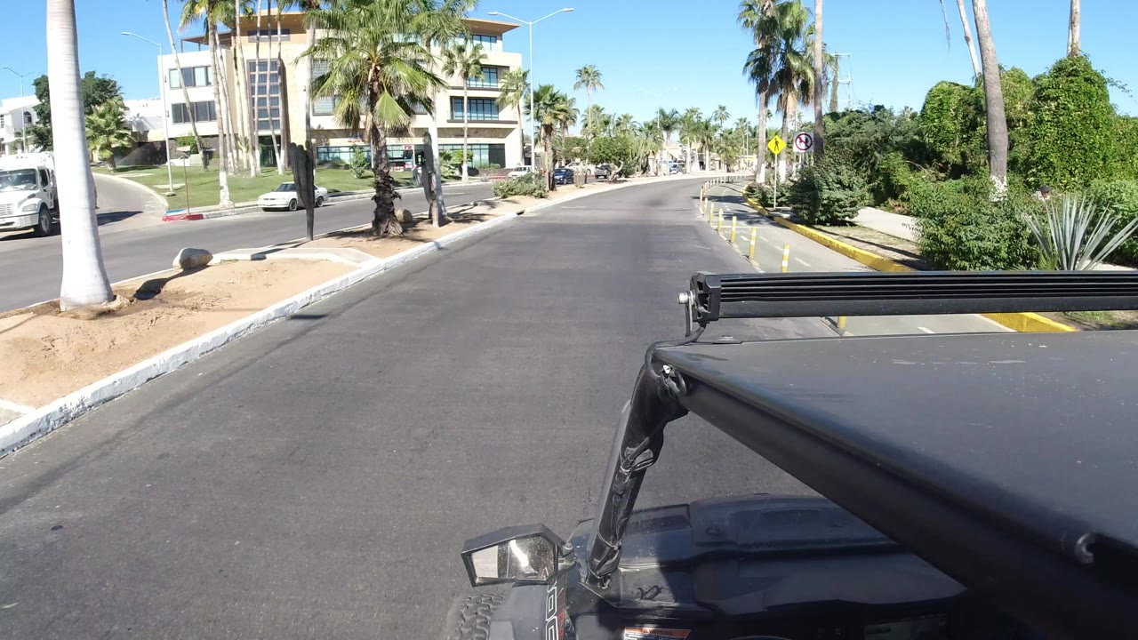 At 0:01, the Los Cabos street remains centered ahead with the Jeep hardware in the lower frame, palms and desert plants on both sides, and a modern building at the left corner.