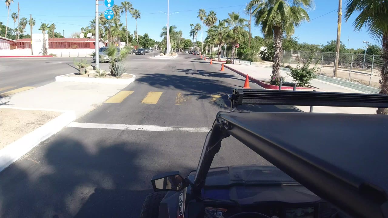 Vehicle-mounted view in Los Cabos, Mexico, looking toward a sunlit street corner with palm trees, cones, sidewalks, parked cars, and no visible people.