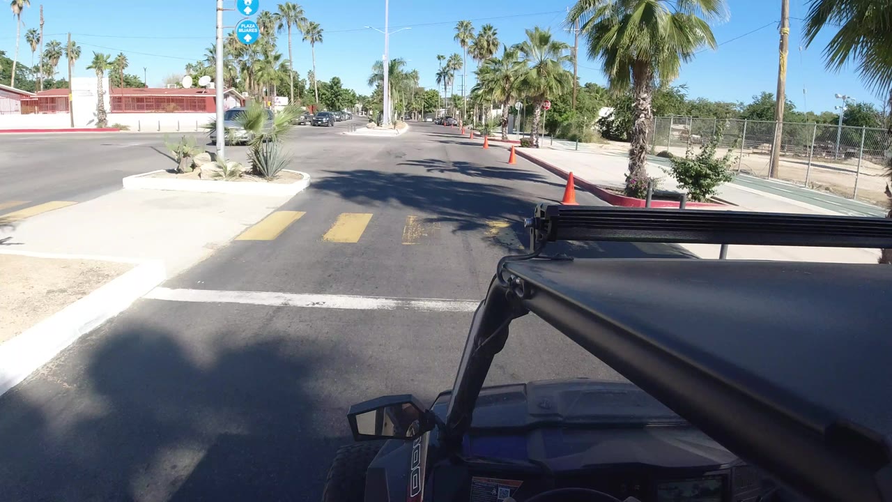 Los Cabos street view from a Wrangler-style vehicle, with black foreground bars, orange cones along the curb, palms, low walls, and no visible people.