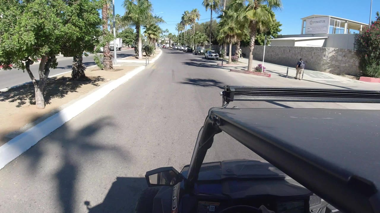 Los Cabos city road seen from a Jeep with palms, parked cars, a low wall, and bright midday shadows at the start of the clip.