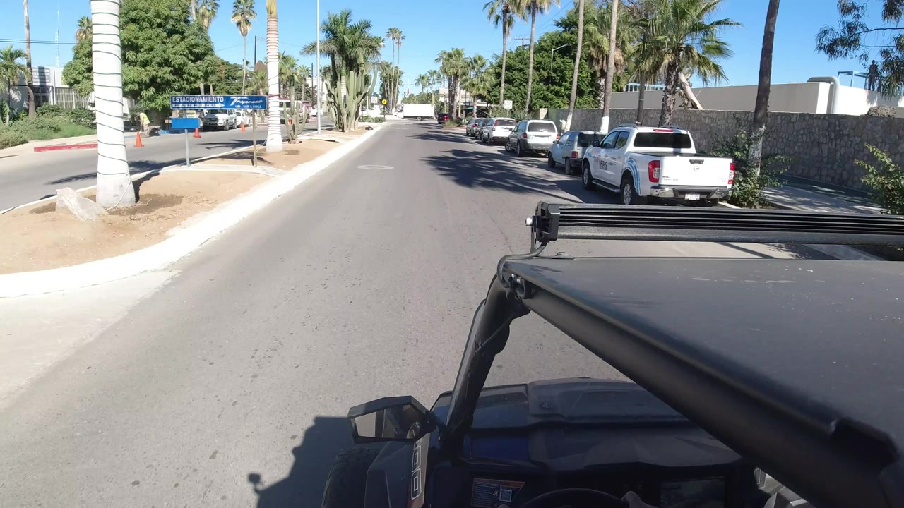 The Wrangler foreground continues down the Los Cabos street, with white parked vehicles at the curb, palms and desert landscaping ahead, and no visible people.
