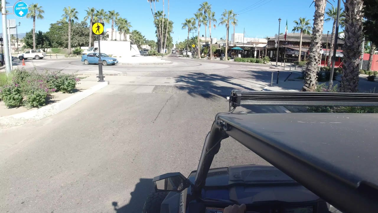 Vehicle POV in Los Cabos with a white box truck and blue car crossing the intersection near palm trees, low buildings, and no visible people.