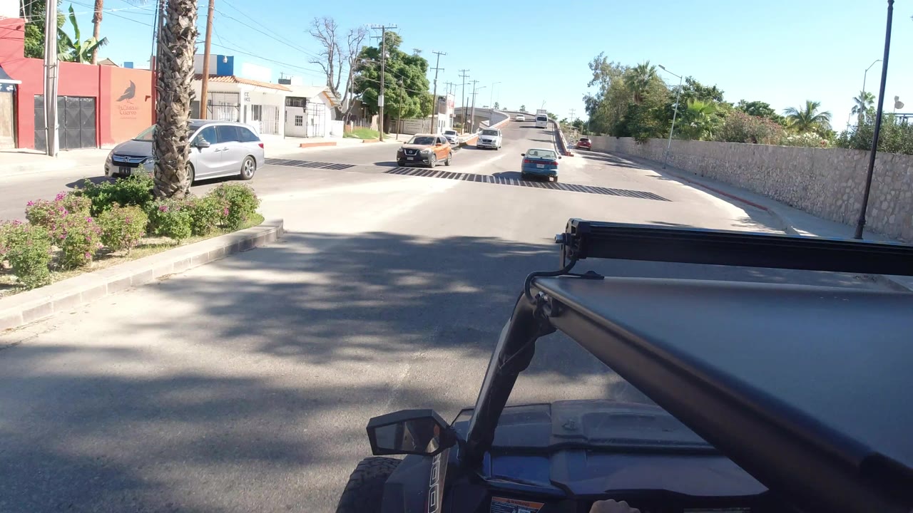 The Jeep roof stays fixed in the foreground as the Los Cabos road continues ahead with a silver car near the curb, distant traffic, roadside trees, and no visible people.
