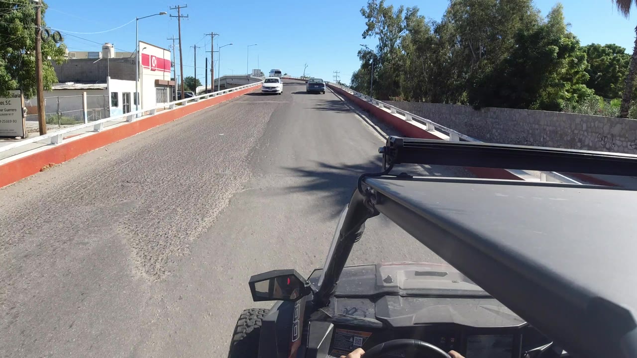 Vehicle-mounted view along the same Los Cabos road, with the bridge railing on both sides, a white car ahead, a roadside commercial building, green trees, and no visible people.