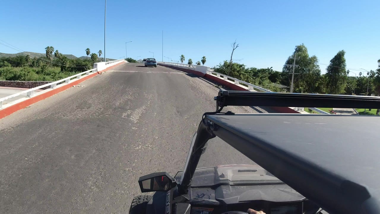 Forward view from a Jeep in Los Cabos, Mexico, approaching a bridge with white rails, red curb trim, green roadside vegetation, low hills, and no visible people.