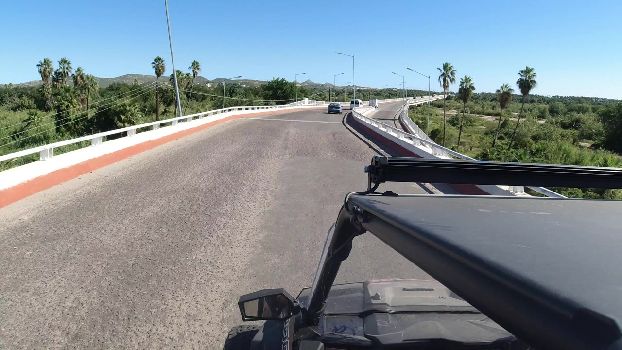 Forward view from a Wrangler on a sunlit Los Cabos road, with white bridge rails, green vegetation, distant hills, a blue sky, and no visible people.