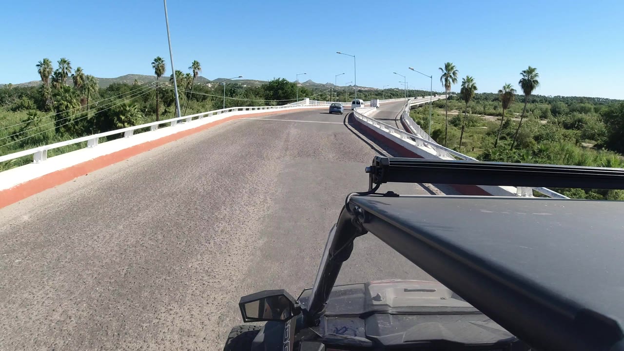 A forward-facing Los Cabos highway view shows the Wrangler frame at bottom right, white guardrails on both sides, palm trees, light traffic, and no visible people.