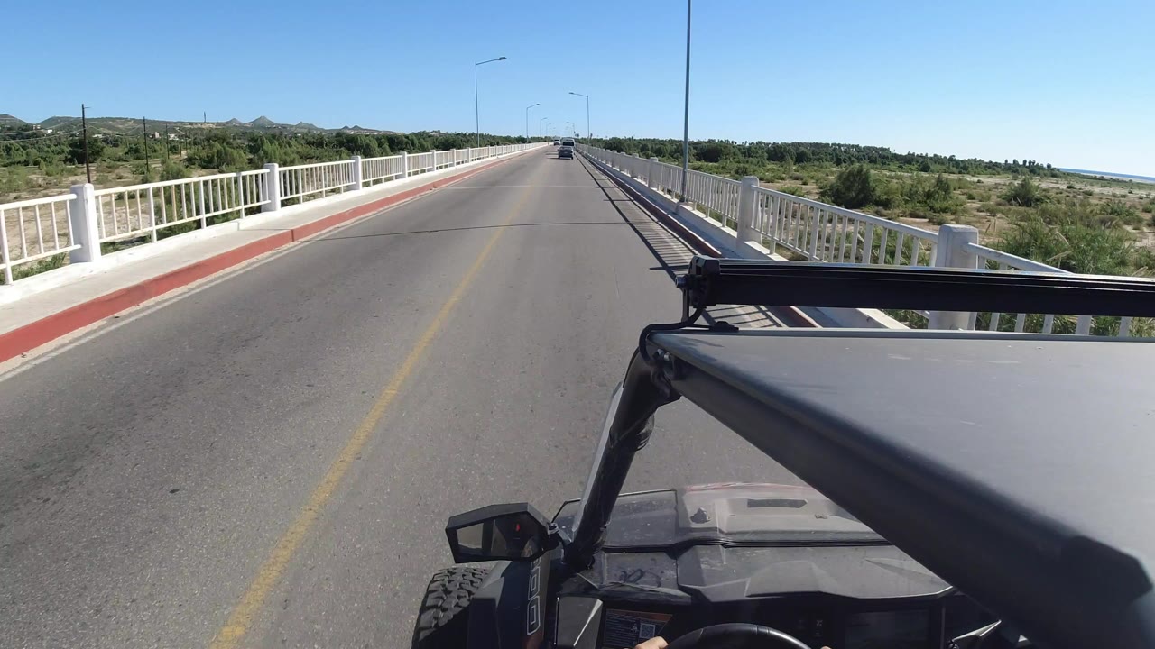 Opening frame of a Los Cabos bridge road seen from a vehicle, with white side railings, green vegetation, low hills, blue sky, and no visible faces.