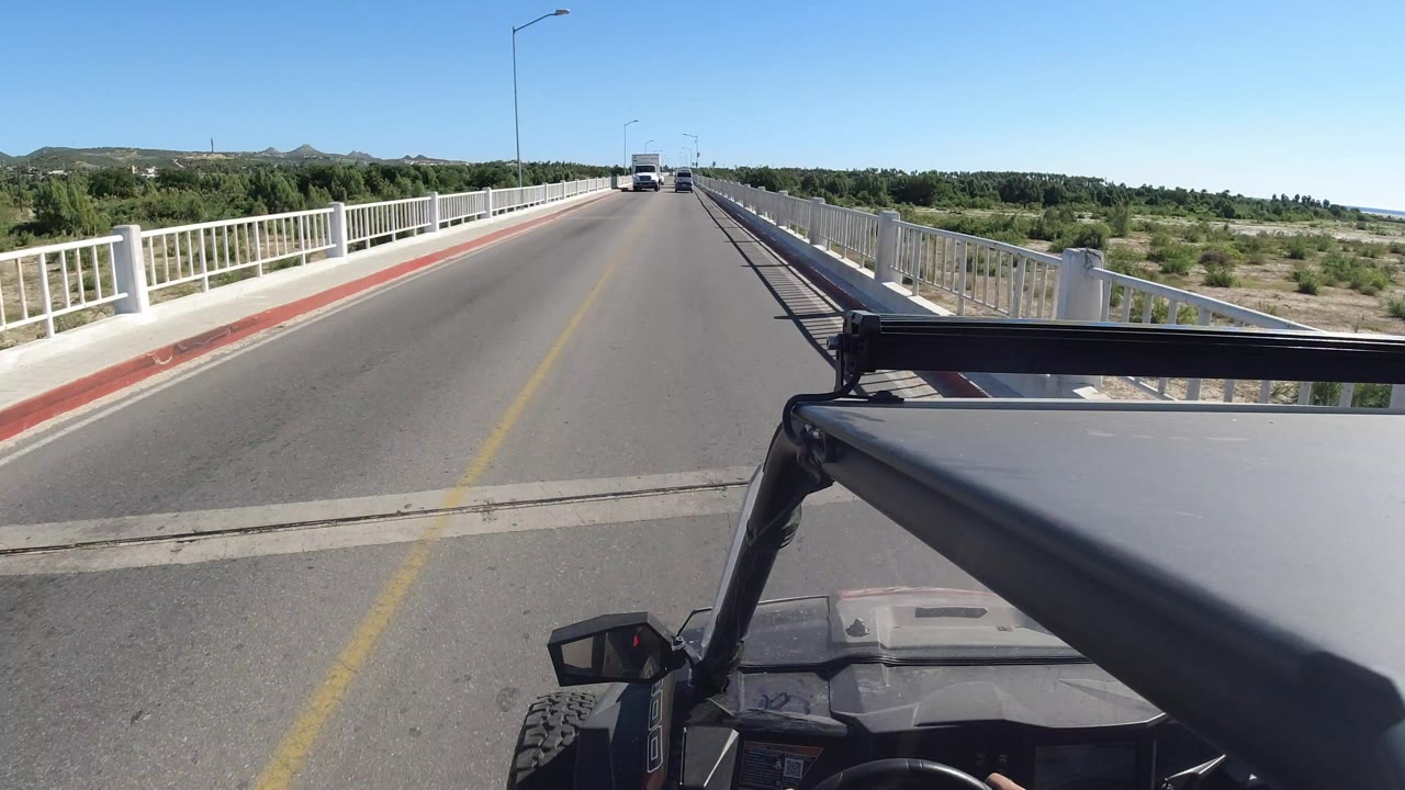 Start of a Los Cabos, Mexico driving view from a Wrangler, with the hood and roll bar in the foreground, white bridge railings on both sides, distant vehicles ahead, green trees, low hills, and no visible people.