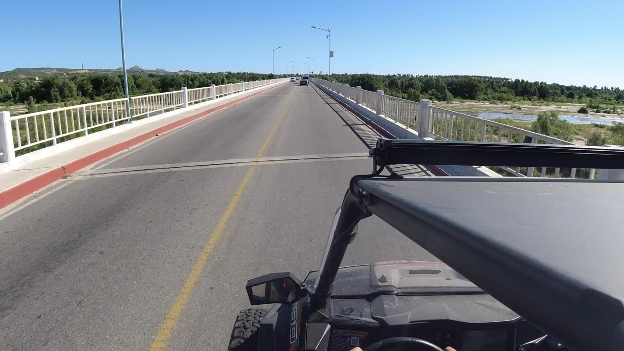 Los Cabos, Mexico driving POV one second in, showing the road centered between white bridge railings, a fixed vehicle foreground, cars in the distance, green vegetation, and no visible faces.