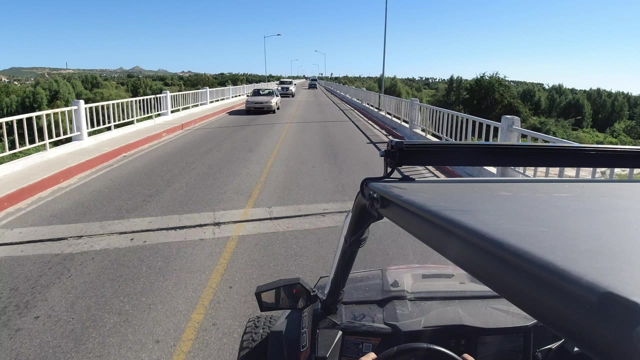 A centered view down the Los Cabos bridge road shows the vehicle hood and roll bar low in frame, railings on both sides, distant cars, and no visible people.