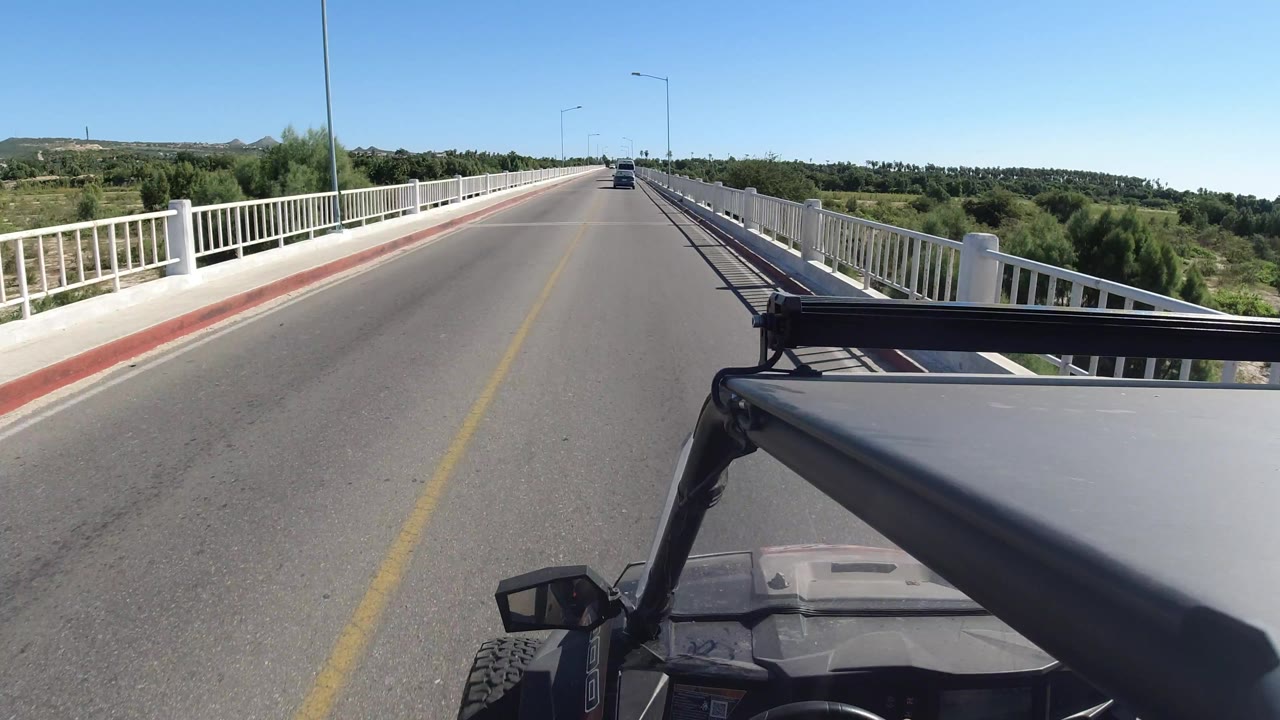 A forward-facing Jeep roof view in Los Cabos, Mexico, shows the road continuing across the bridge with white railings, low greenery, a distant car, and no visible people.