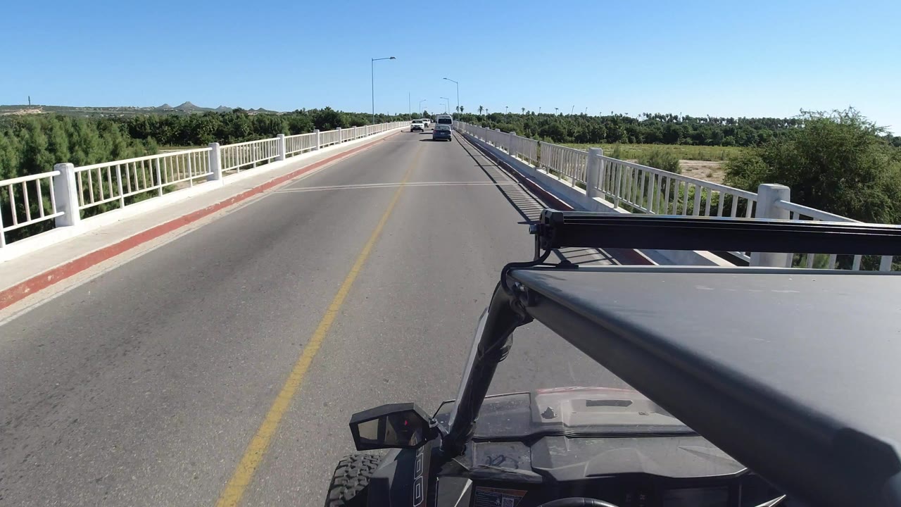 Opening Los Cabos frame from a jeep perspective on a two-way bridge, with the hood and roll bar in the foreground, white railings on both sides, distant cars ahead, green vegetation beyond the bridge, and no visible people.