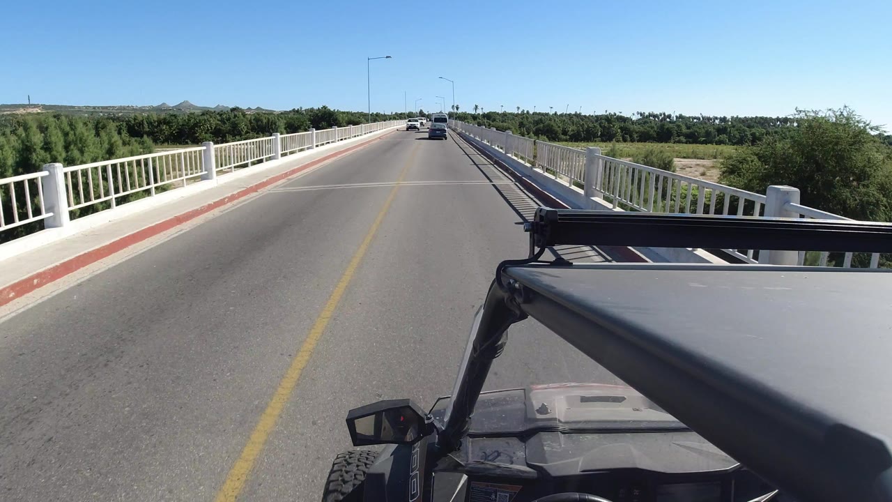 Los Cabos bridge view one second in, with the jeep hardware steady at the lower right, a yellow center line running forward, white guardrails, trees beyond the bridge, small cars ahead, and no visible people.