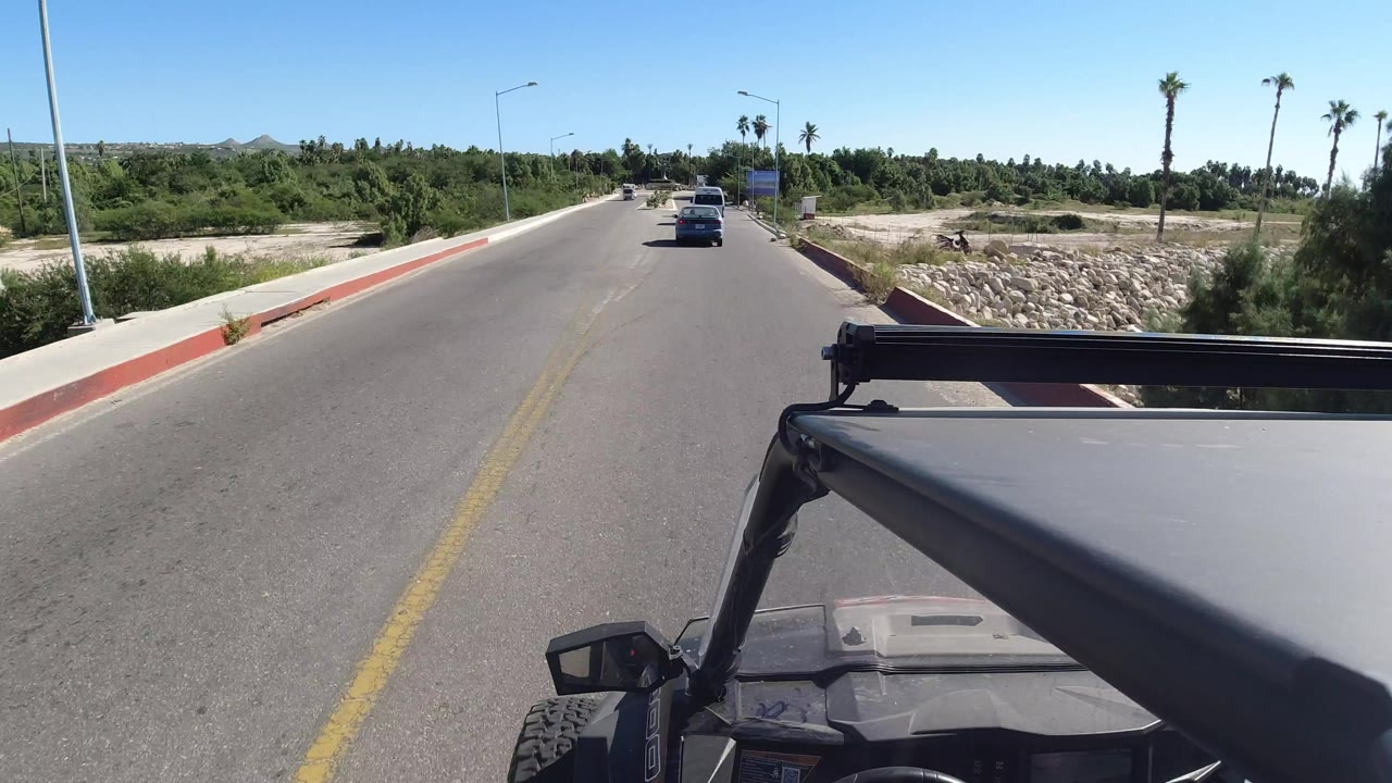 Opening view from a Wrangler on a Los Cabos two-lane road, with the roof and roll bar in the foreground, cars ahead, palms, dry roadside ground, and no visible people.