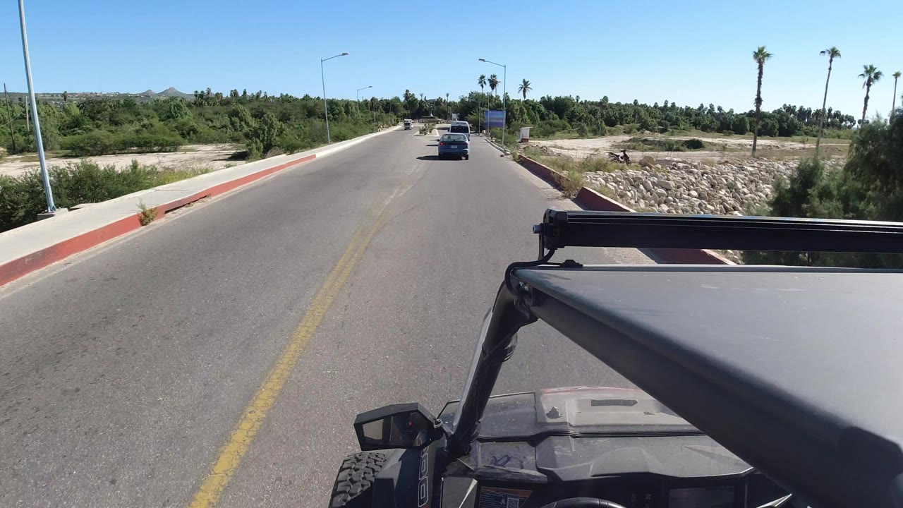 A slightly later Los Cabos driving frame shows the Wrangler roof lower right, the road stretching forward, a car in the lane ahead, palms, roadside stones, and no visible people.