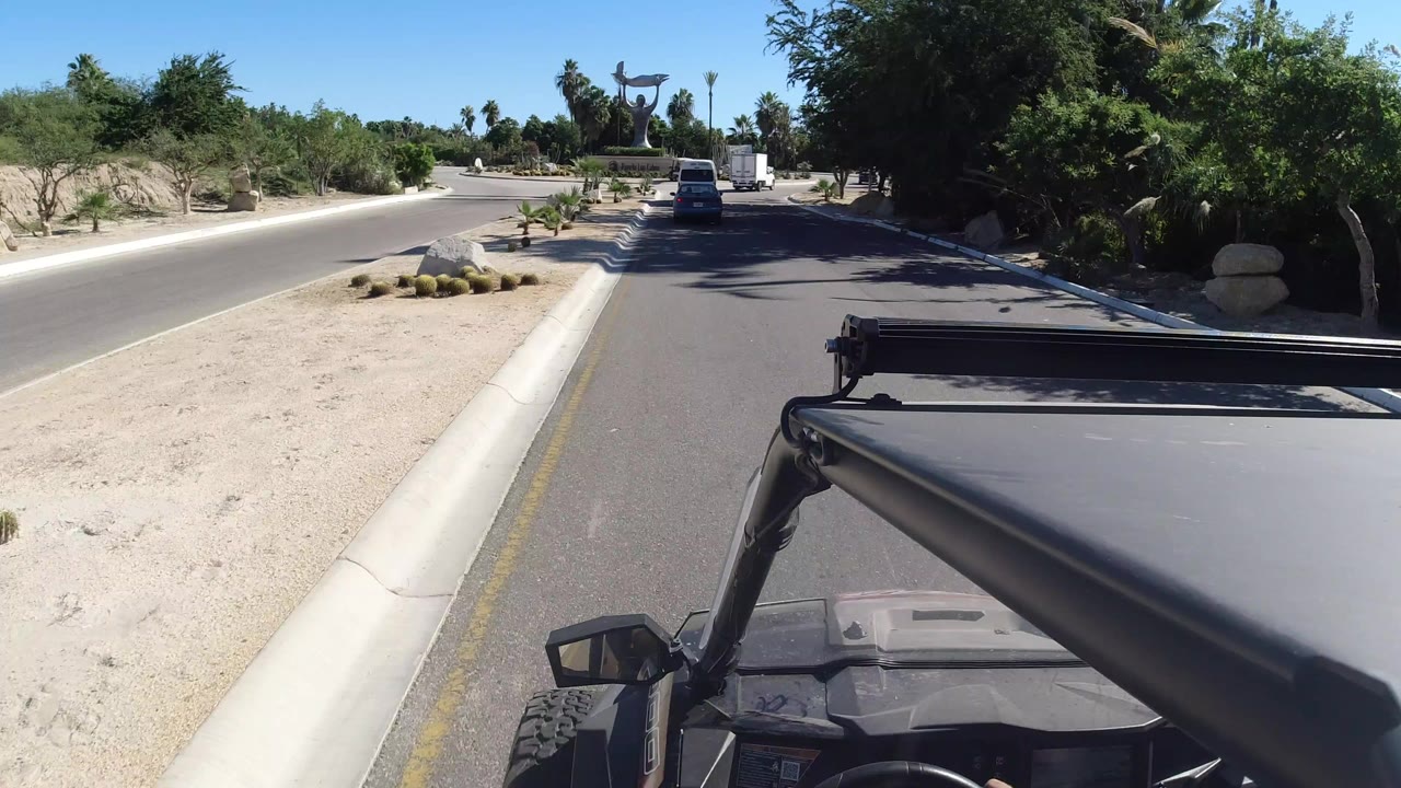 A Los Cabos road seen from an open vehicle, with desert gravel, palms, a white vehicle ahead, a large sculpture in the distance, and no clearly visible people.