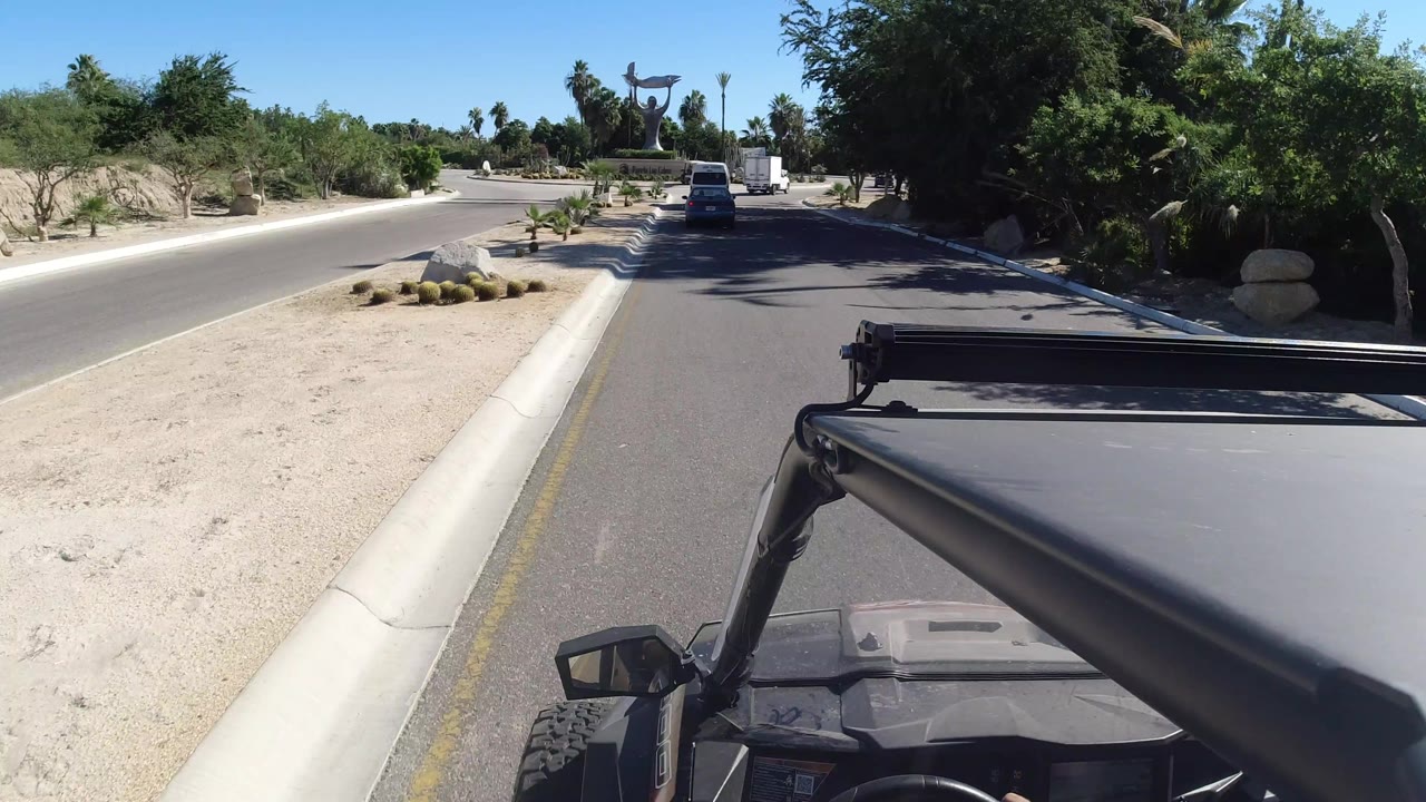 A forward road view in Los Cabos with the open vehicle frame in the foreground, a white vehicle ahead, pale gravel landscaping, palms, and the distant roadside sculpture.