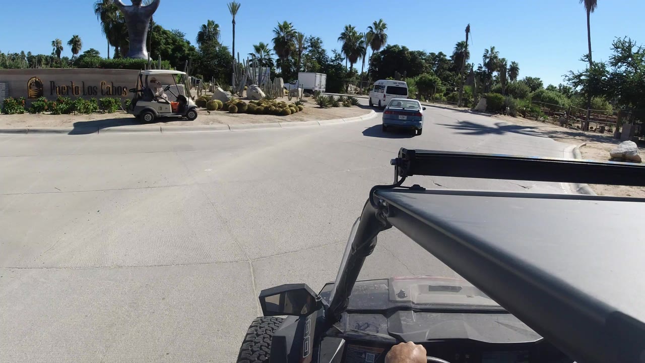 Forward vehicle POV in Los Cabos with a black roll bar in the foreground, a roadside entrance sign, palms, cactus landscaping, cars ahead, and no visible faces.