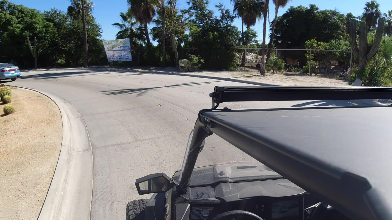 Start of the Los Cabos driving clip with the black jeep roof and light bar in the foreground, a curved paved road ahead, palms and dense trees beyond, and no visible people.