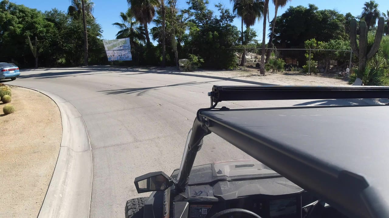 Los Cabos road view one second in, with the jeep roof still filling the lower frame, a sunlit curb and road ahead, dense greenery, palms, and no visible people.